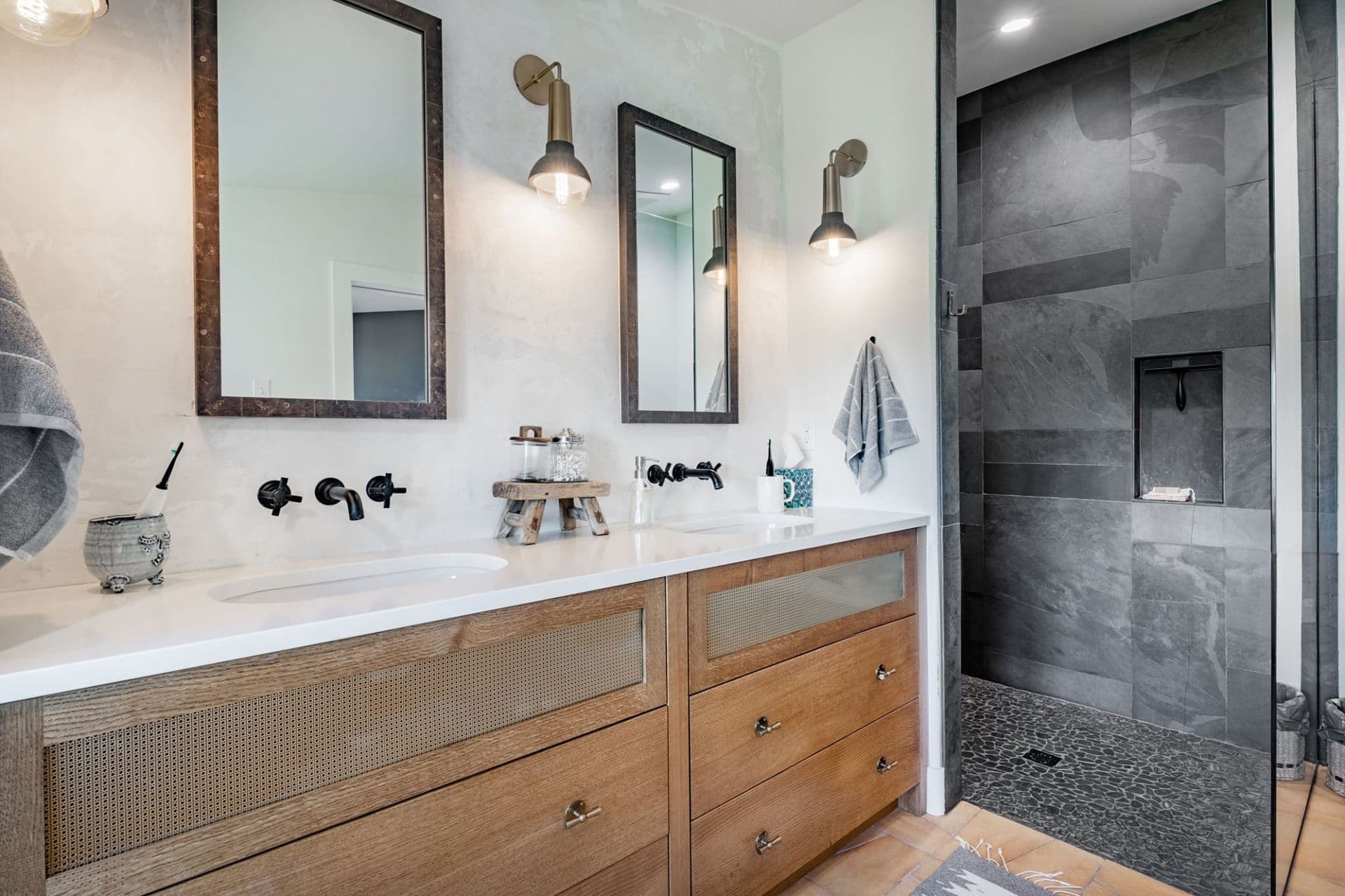 Modern Farmhouse style bathroom with dual vanity featuring white countertop and wood cabinetry, two framed mirrors, brass sconces, black wall-mounted faucet, and walk-in shower with gray tile and glass enclosure. Total Bath Systems