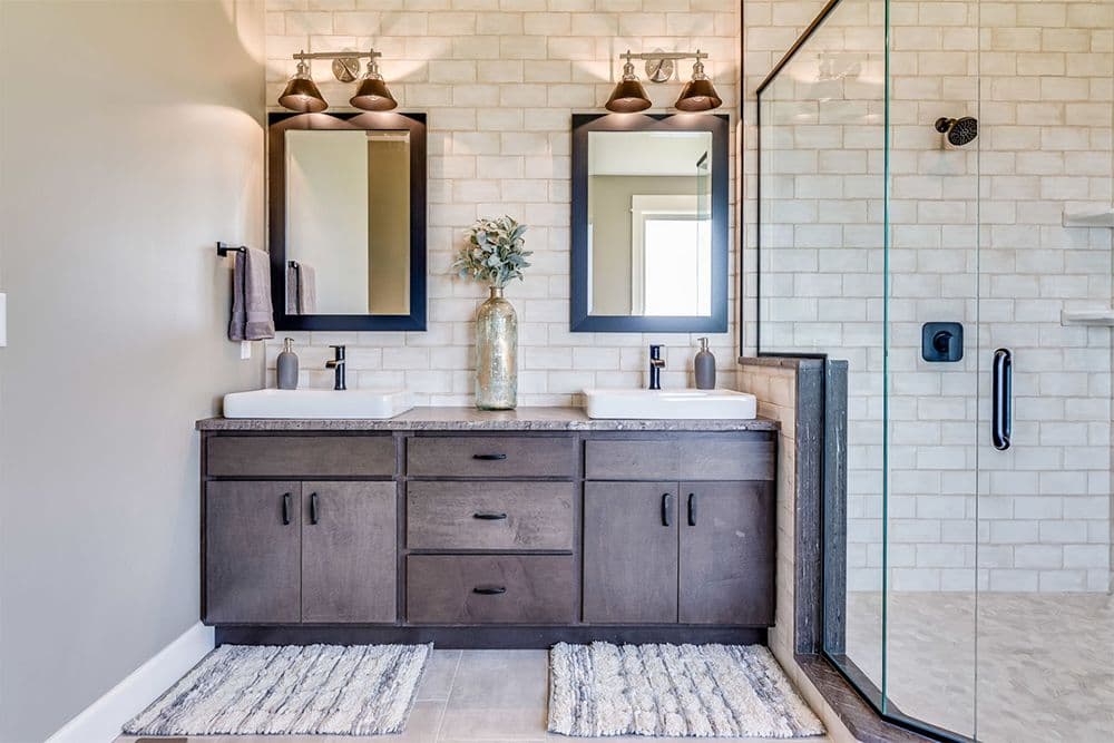 Modern Farmhouse style master bathroom with dual vanity, two framed mirrors with bronze sconces, subway tile walls, walk-in shower with glass enclosure, and decorative vases