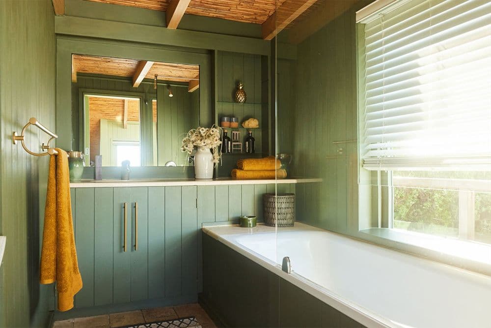 Modern farmhouse bathroom with green shiplap walls, white soaking tub by window, floating vanity with mirror, and wooden beam ceiling details