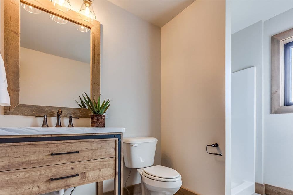 Modern Farmhouse Style bbathroom with wood vanity, white sink countertop, framed mirror with overhead lighting, potted plant, white toilet, and doorway to adjoining room