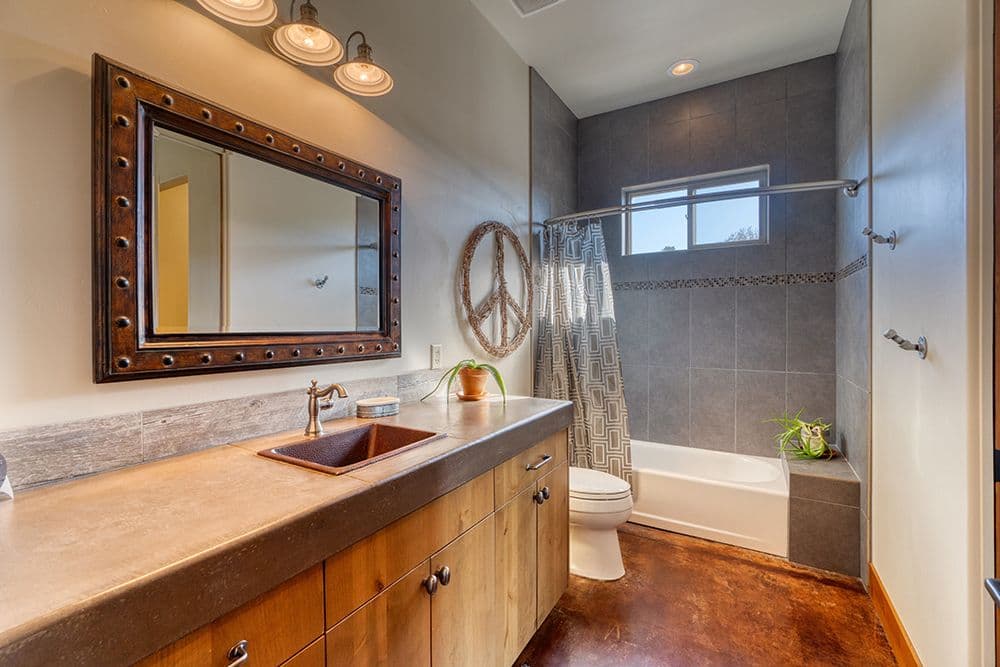 Modern Farmhouse bathroom with wooden vanity, copper sink, decorative mirror with studded frame, walk-in shower with gray tile, and peace sign wall decor. Total BaTH Systems