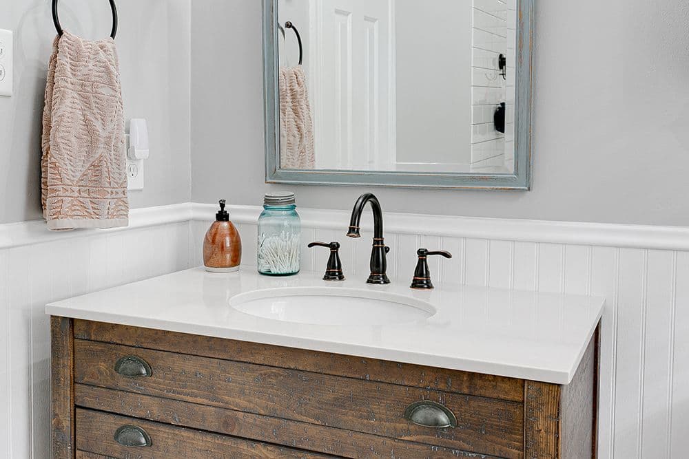 Bathroom vanity with white countertop and undermount sink, bronze faucet, wooden dresser base, framed mirror, and decorative towel on hook