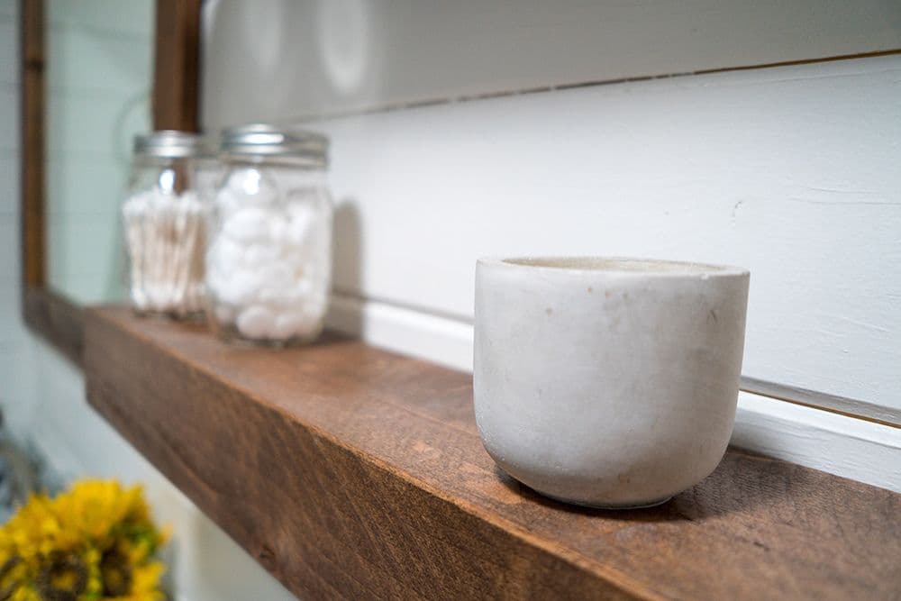 Modern Farmhouse style bathroom shelf of rough wood with concrete planter, glass jars holding cotton swabs and rounds, and yellow flower accent