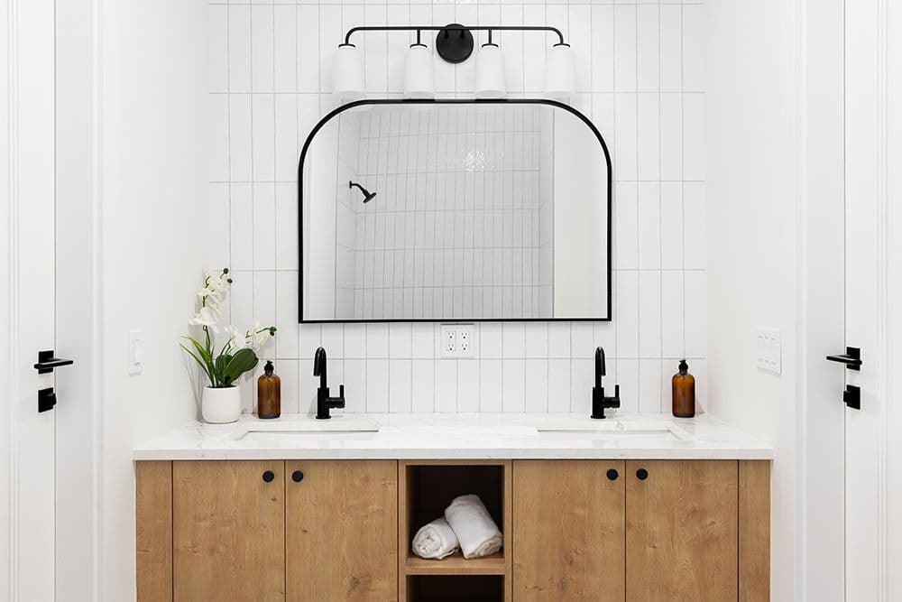 Modern Farmhouse bathroom vanity with white subway tile wall, arched black-framed mirror, dual black faucets, potted orchid, amber bottles, and natural wood cabinetry with towel storage. Total Bath Systems